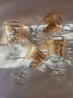 Four clear glass goblets with gold detailing and gold-trimmed rims, arranged on a sunlit ivory surface with dramatic light reflections.