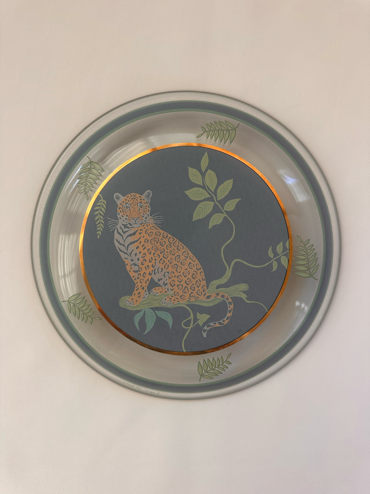 A round decorative plate featuring a central leopard illustration on a dark gray background, surrounded by green leaves and a gold inner ring, with a gray outer rim.