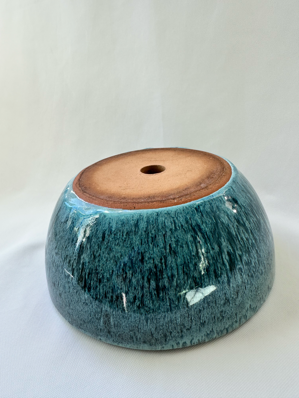 A handmade ceramic planter or bowl with a glossy teal-blue glaze and a natural brown clay base, shown on a white fabric background.