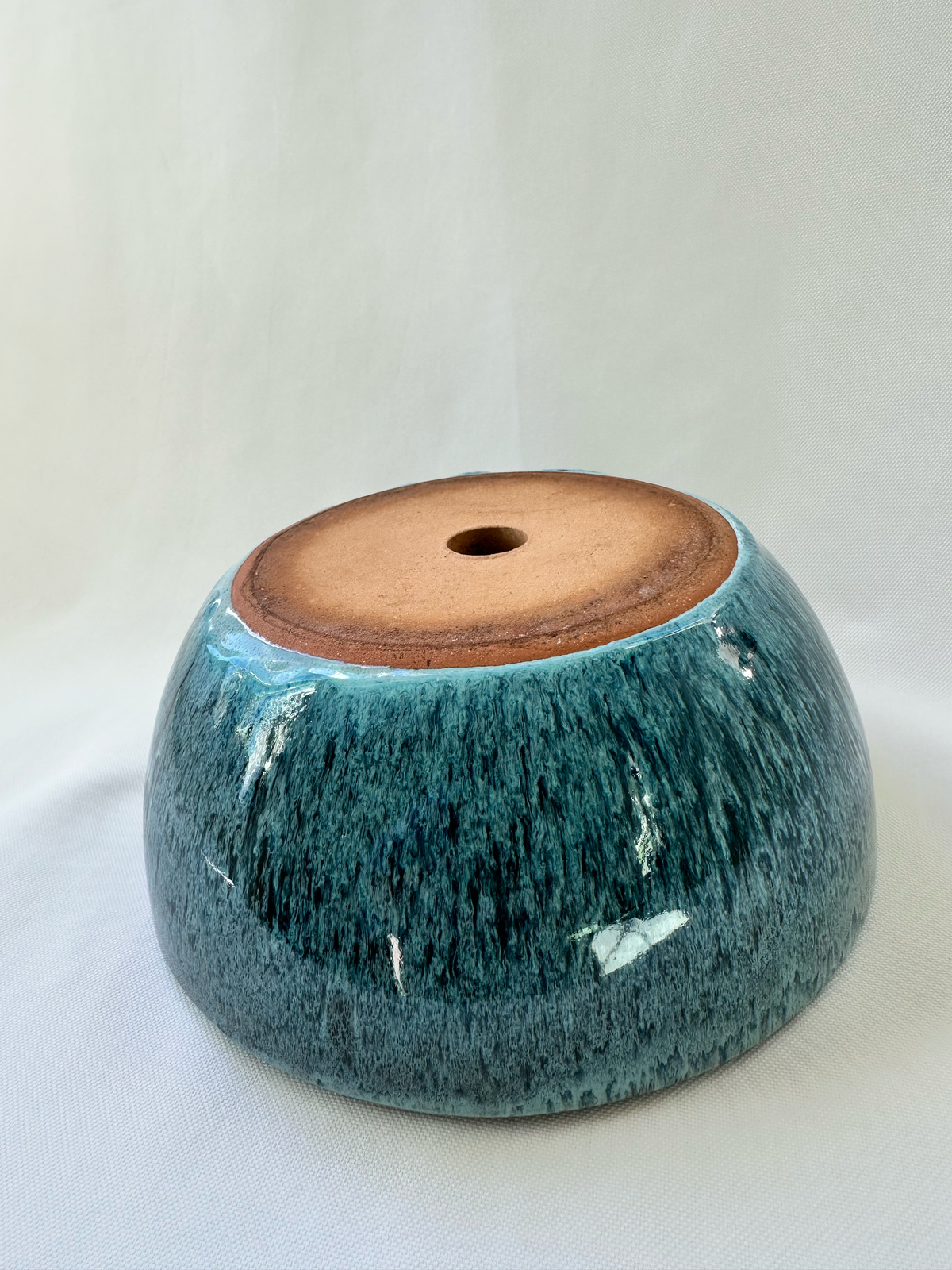 A handmade ceramic planter or bowl with a glossy teal-blue glaze and a natural brown clay base, shown on a white fabric background.