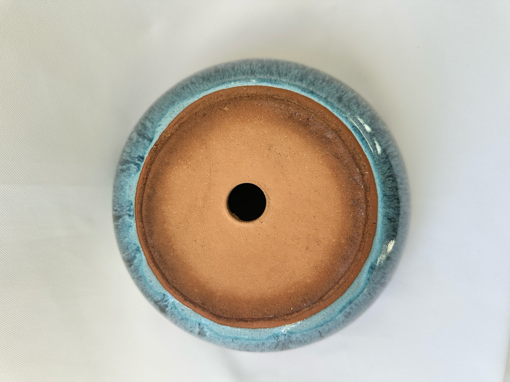 Bottom view of a round ceramic pot base with a glossy light blue glazed rim and a matte terracotta-colored center, featuring a single drainage hole.