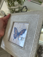 A hand holds a square silver glitter picture frame with beaded borders and a butterfly print, shown in a home setting.