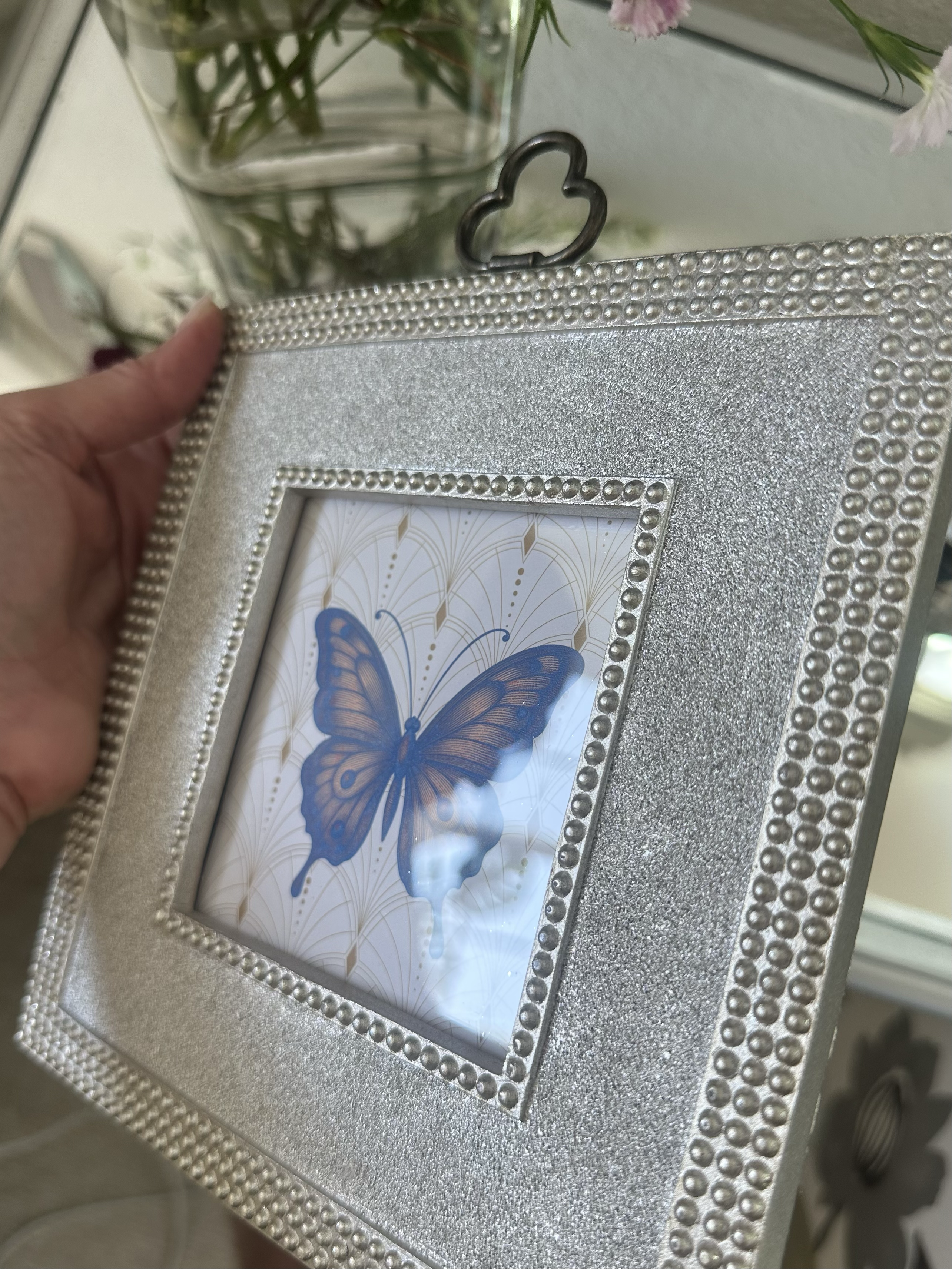 A hand holds a square silver glitter picture frame with beaded borders and a butterfly print, shown in a home setting.