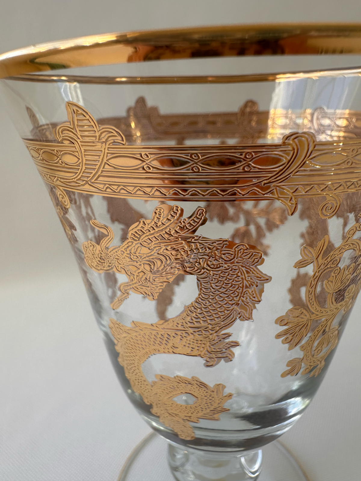 A close-up of a clear glass goblet or tumbler with ornate gold dragon and scroll motifs etched and hand-painted across the body, plus a wide gold rim, shown against a plain light background.