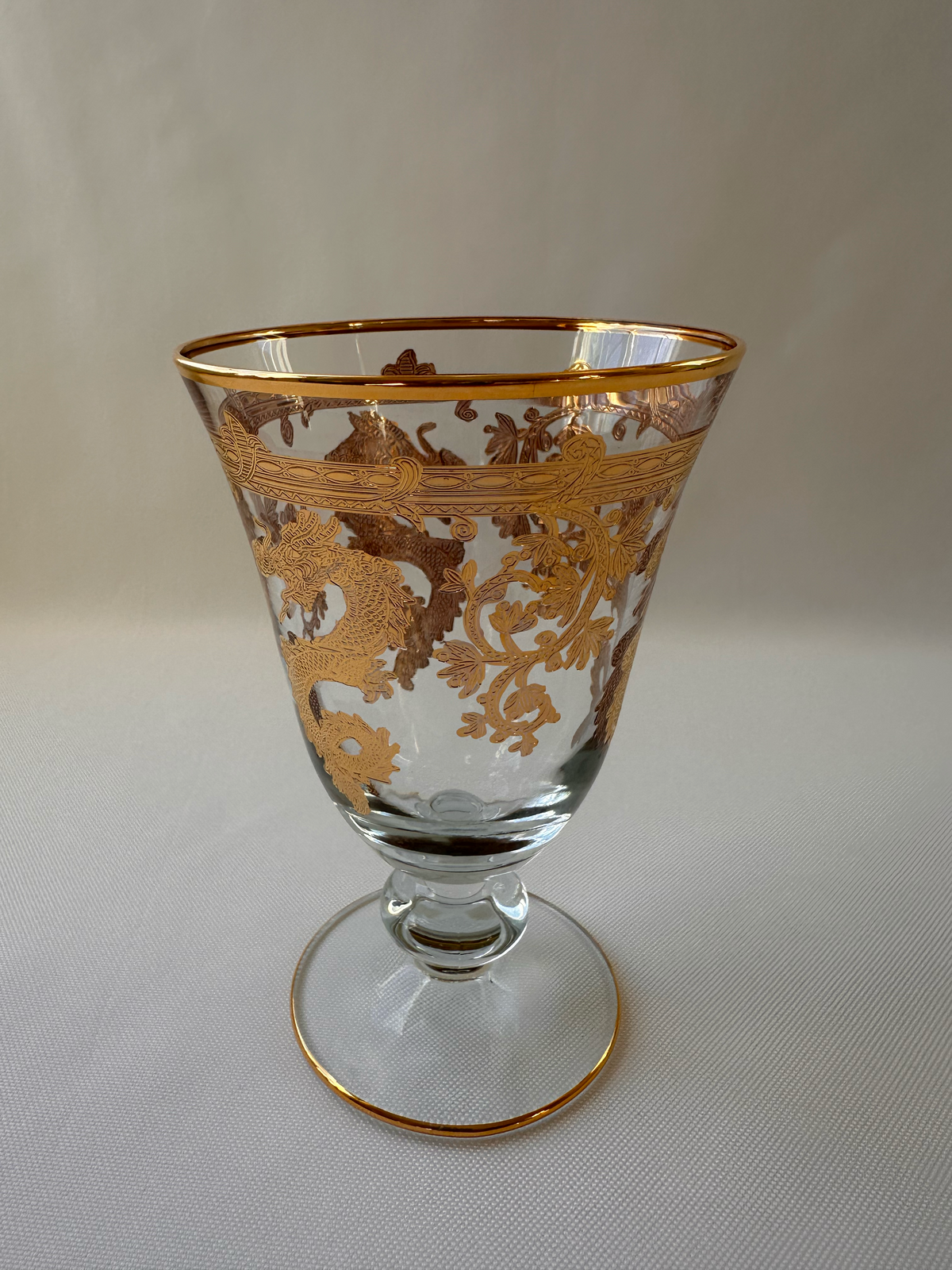 A clear glass goblet with ornate gold floral patterns and gold trim, shown on a light gray textured background.
