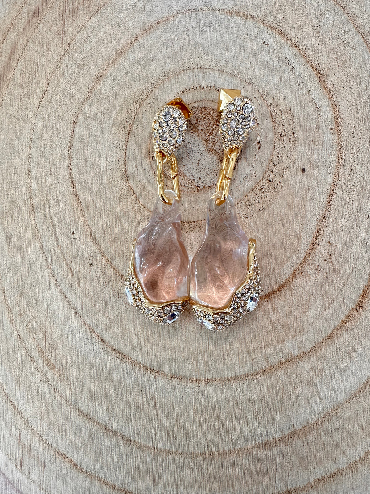 Close view of Alexis Bittar clear Liquid Lucite drop earrings with gold-tone links and crystal pavé accents on a textured neutral surface.
