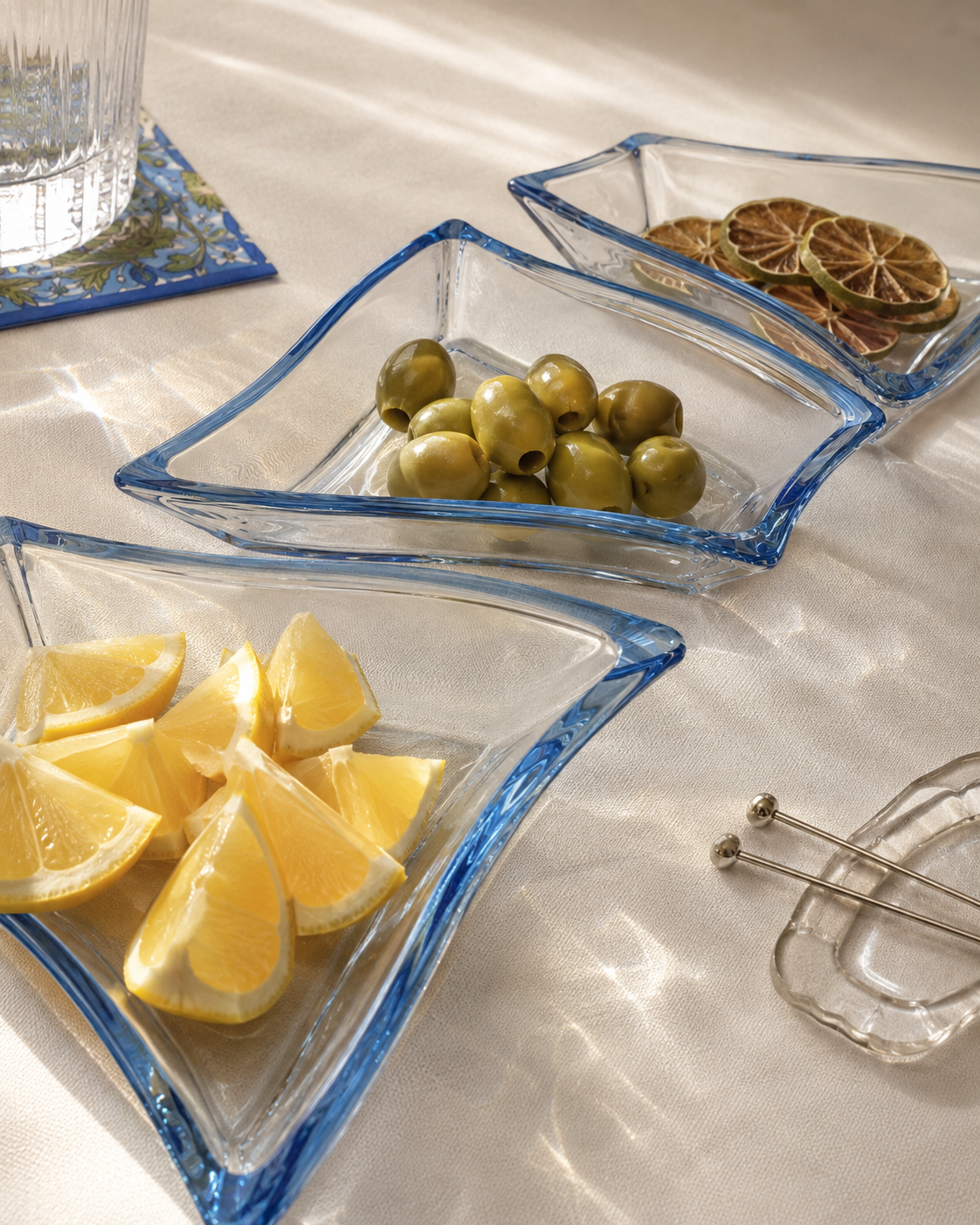 3 clear bowls with blue rims sit on a cocktail table holding olives, fresh lemon wedges and dried lime wheels. 