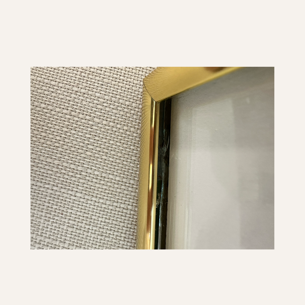 Close-up of a gold-framed picture frame against a textured beige fabric background revealing slight cracking of glass beneath the frame.
