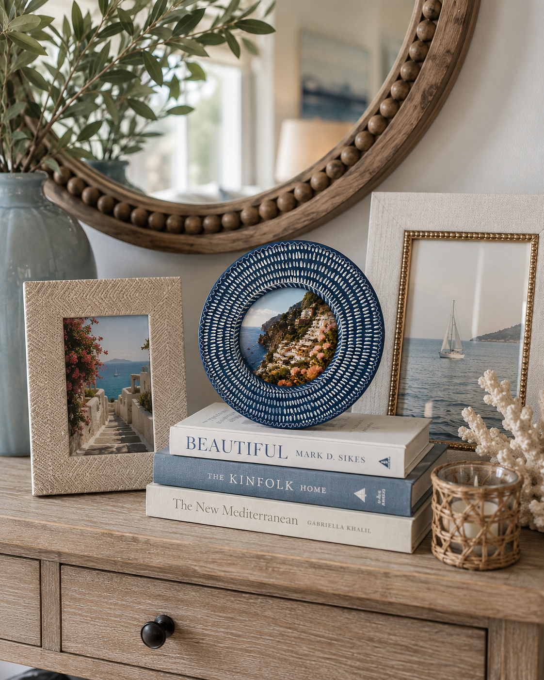 A styled interior scene showing a round blue and white patterned decorative plate on a stack of books, alongside two picture frames, a mirror, and other home decor on a wooden console table.