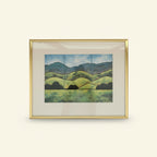 A framed landscape art print featuring green rolling hills, dark tree silhouettes, and blue mountains under a cloudy sky, enclosed in a gold frame with a white mat.