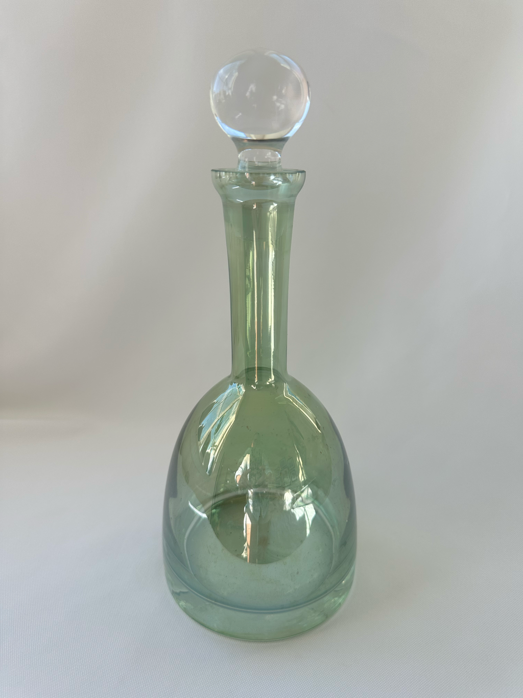 Full view of pale green iridescent glass decanter with tall neck and clear globe stopper.
