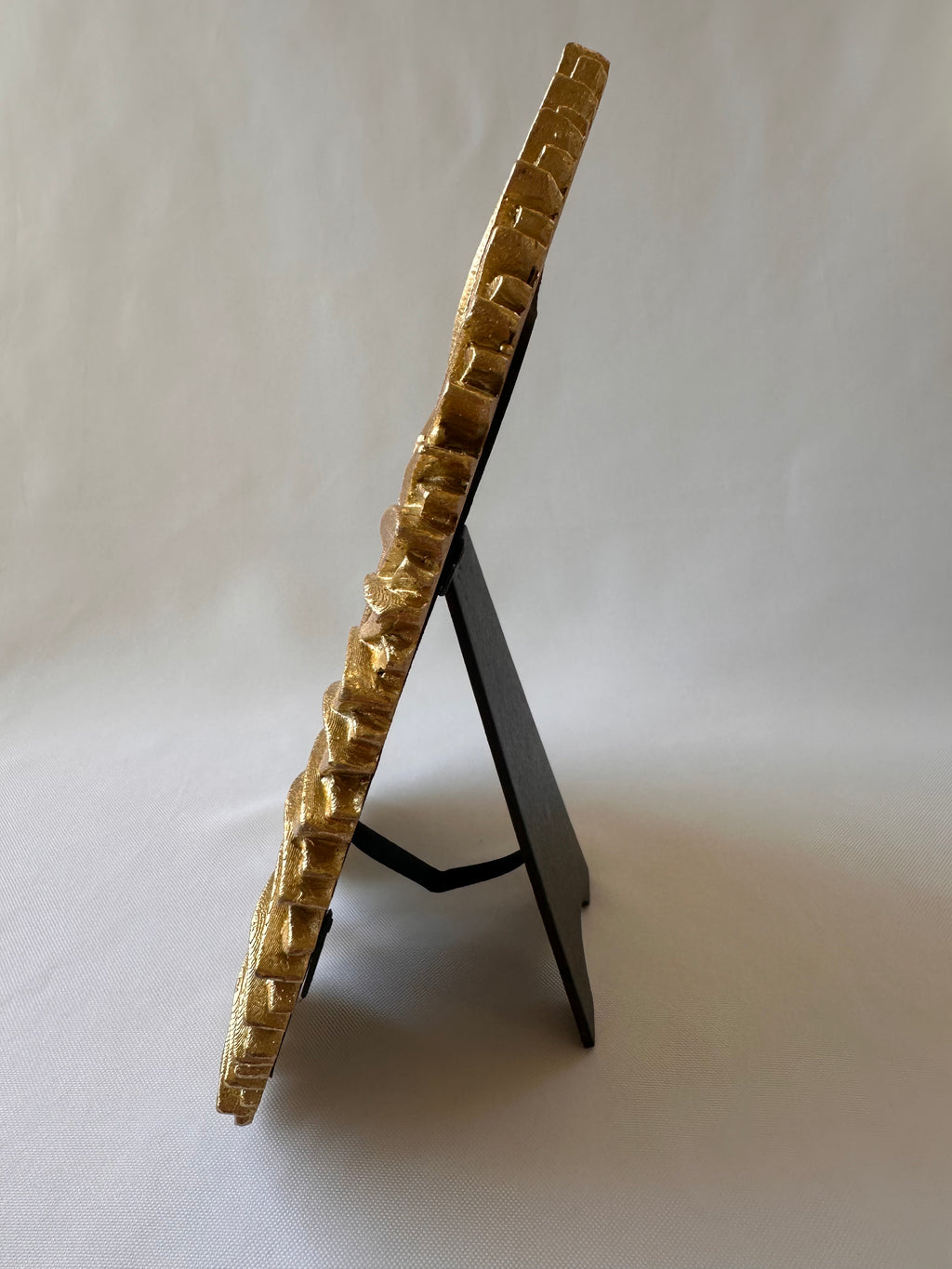 A gold-framed picture frame with a textured, geometric patterned border, displayed standing on a black easel against a light gray background.