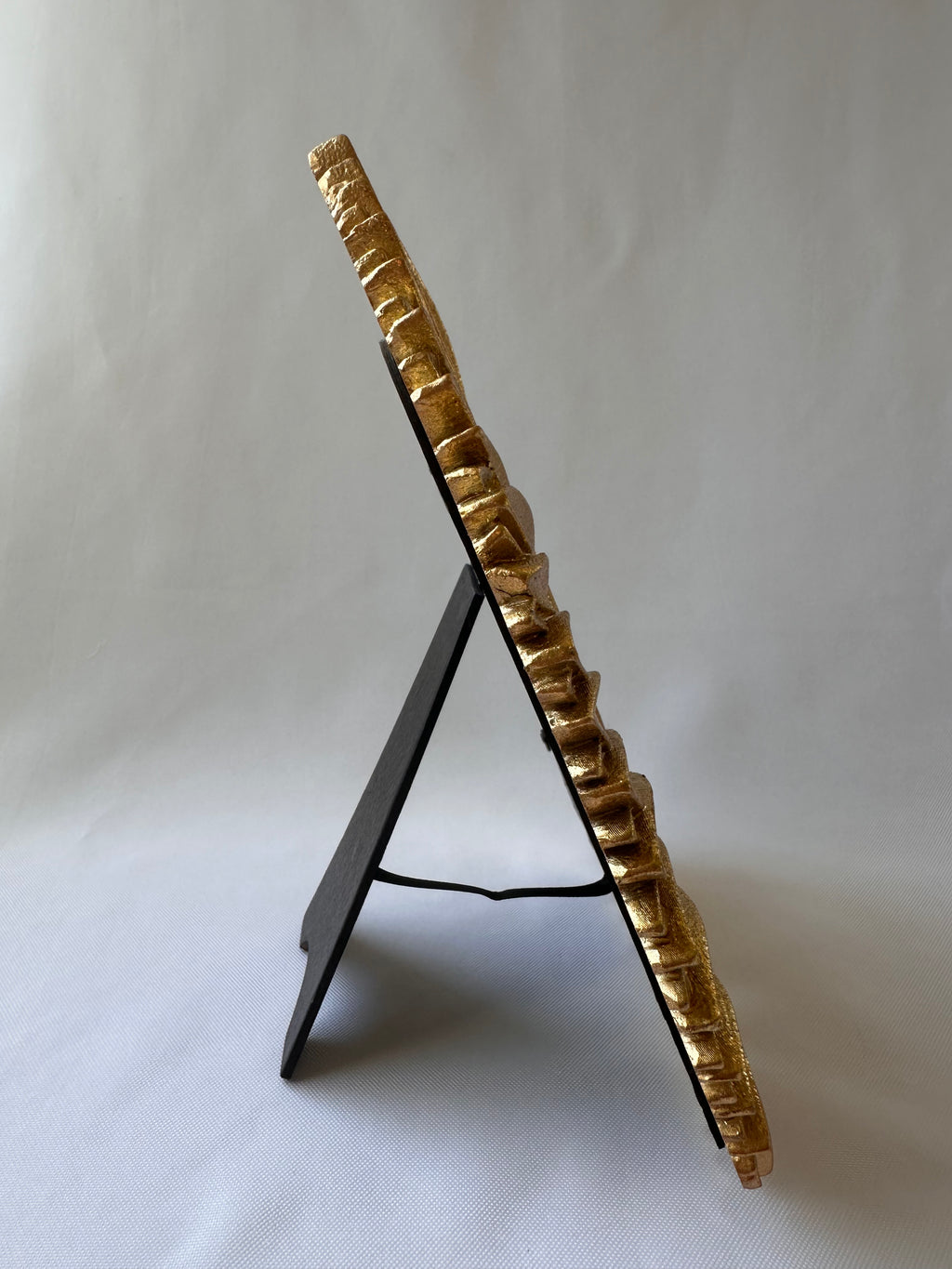 A gold-framed picture frame with a textured, ribbed border, displayed on a black A-frame stand against a light gray fabric backdrop.