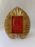 A gold metallic picture frame with a sunburst or fan-like radiating design, shown holding red patterned paper insert. The brand name 'ASHLAND' is visible on the insert.