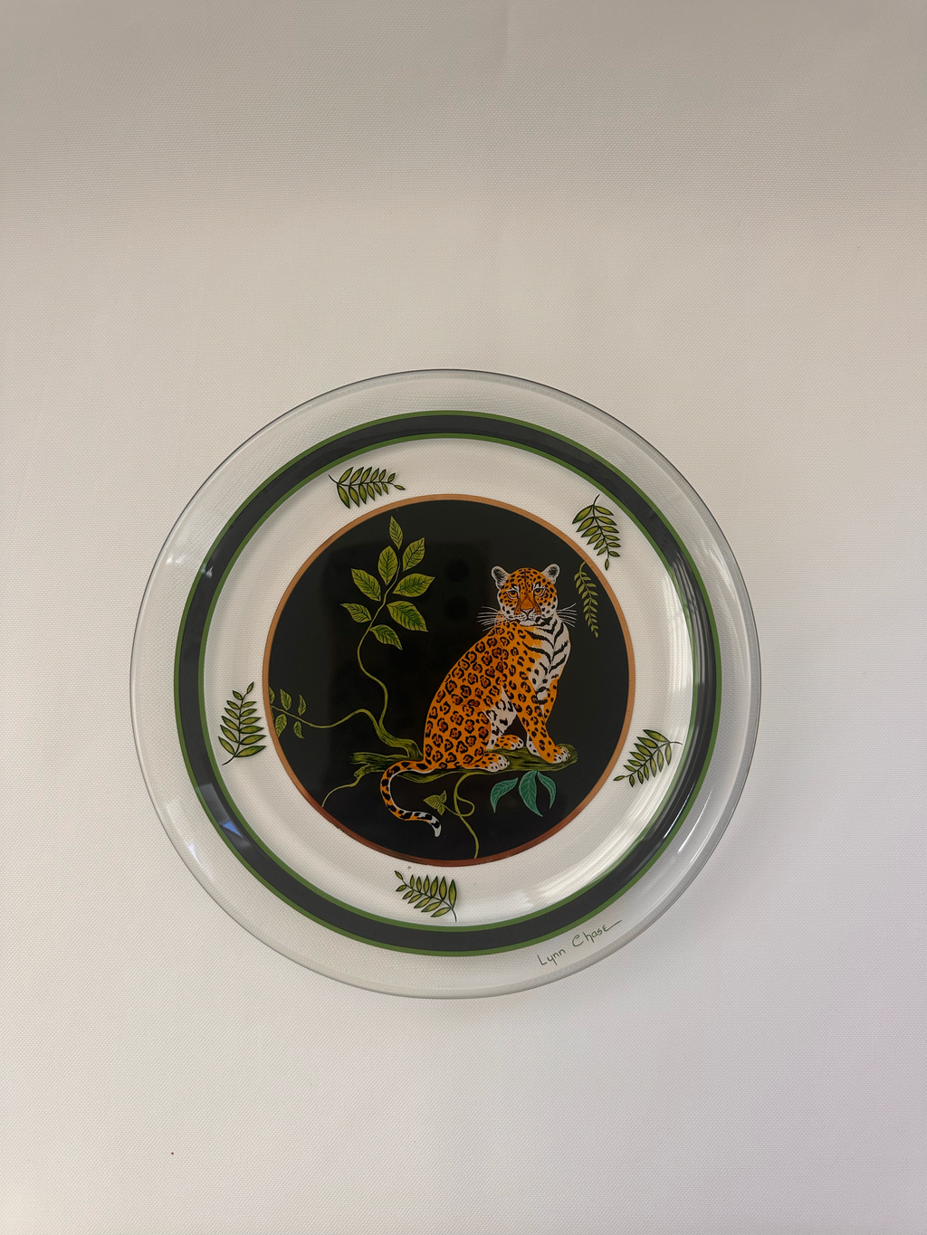 A round decorative plate featuring a central leopard illustration on a black background, surrounded by green leaves and bordered by concentric dark green rings on a white base.