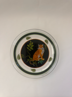 A round decorative plate featuring a central leopard illustration on a black background, surrounded by green leaves and bordered by concentric dark green rings on a white base.