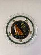 A round clear glass plate featuring a printed design of a leopard on a black background with green leaves and vines, bordered by dark green rings.