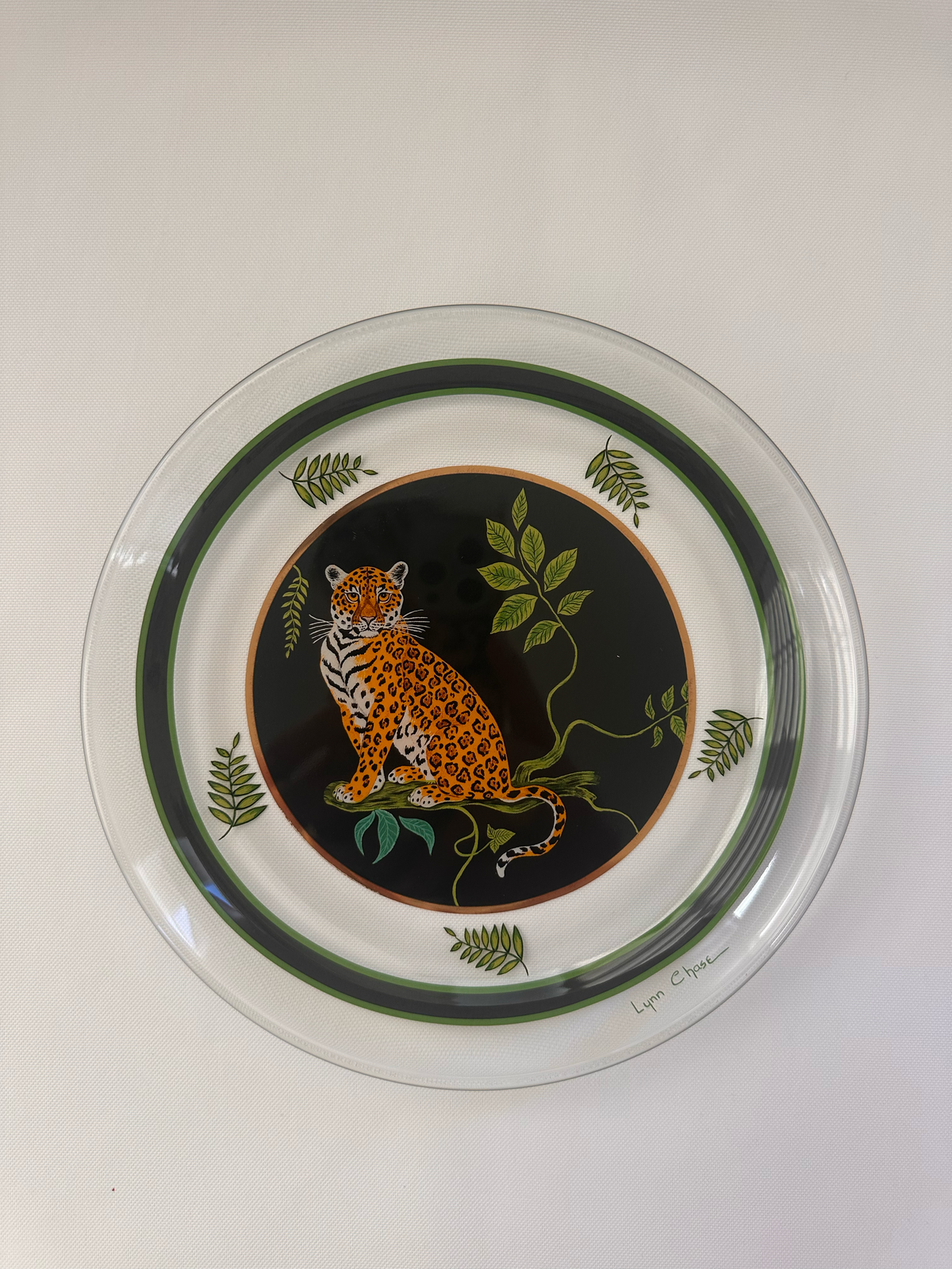 A round clear glass plate featuring a printed design of a leopard on a black background with green leaves and vines, bordered by dark green rings.