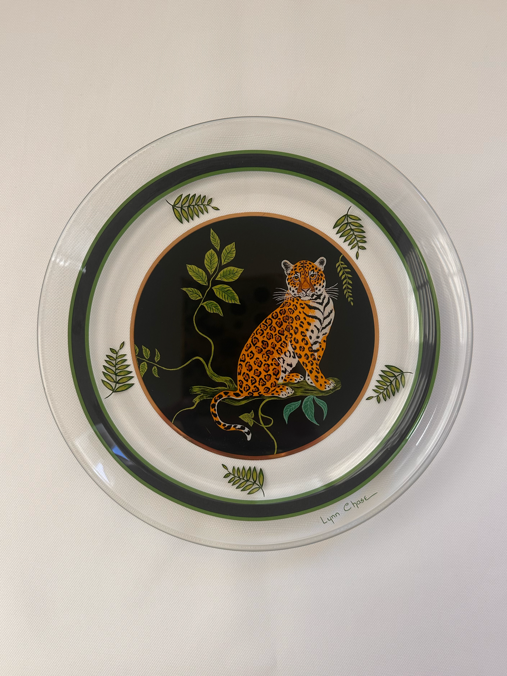 A round white ceramic serving platter with a clear glass or plastic rim, featuring a central black oval with an orange and black leopard, surrounded by green leaves and dark green concentric rings.
