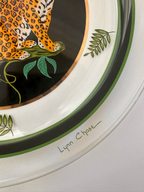 Close-up of a decorative ceramic plate featuring a leopard illustration, green botanical leaves, and a white rim with a green stripe. The name 'Lynn Chase' is printed on the rim.