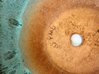 Close-up of a terracotta-colored ceramic sink base with a white drain cover, showing the maker's mark 'MD PMD' stamped on the surface, set against a background of turquoise water.