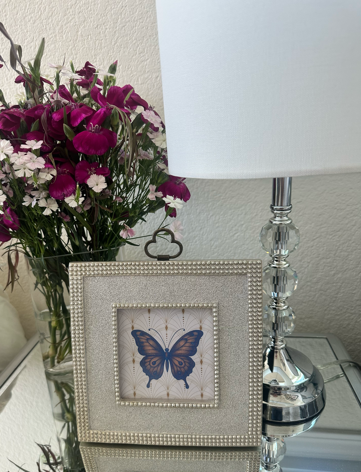 A square framed butterfly art print with a silver beaded frame, styled on a bedside table next to a lamp and a vase of flowers.