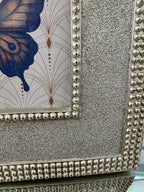Close-up of a decorative framed artwork with a silver beaded border, textured silver surround, and a butterfly motif in blue and gold on a light background.
