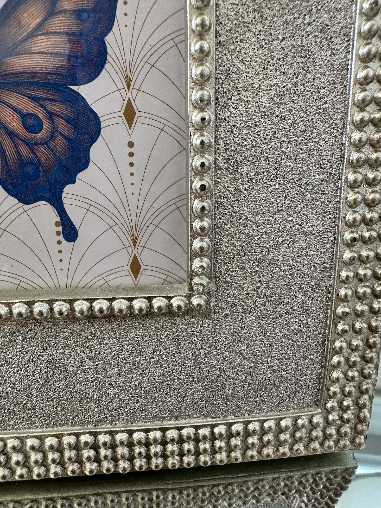 Close-up of a decorative framed artwork with a silver beaded border, textured silver surround, and a butterfly motif in blue and gold on a light background.