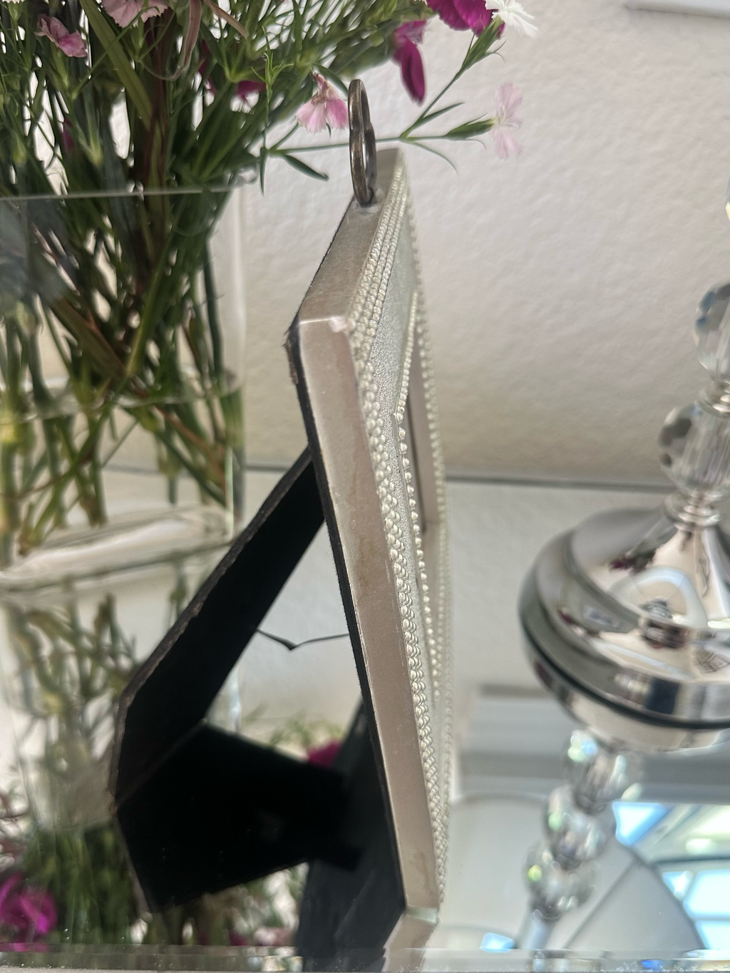Profile of a silver-toned rectangular picture frame with a beaded border and black stand, shown standing on a reflective surface with flowers and a silver candle holder in the background.