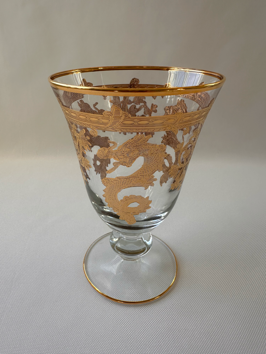 A clear glass goblet with ornate gold dragon motifs and gold trim, shown on a light textured background.