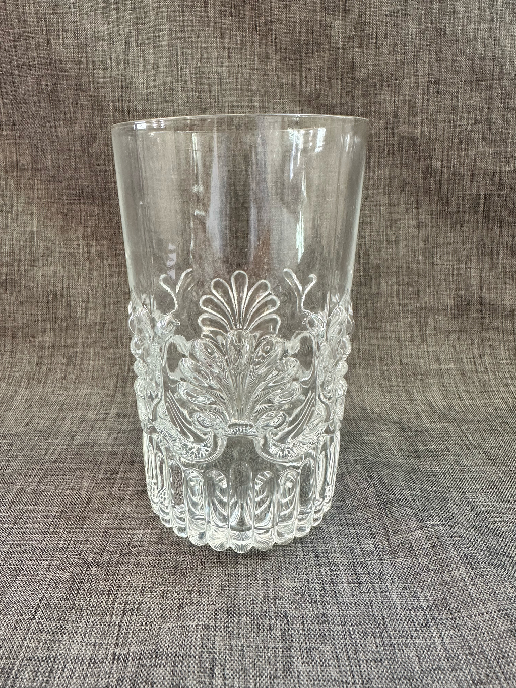 A clear glass tumbler with an embossed floral and scroll pattern, sitting on a textured gray fabric background. Vintage MMA reproduction glasses. 