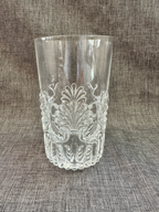 A clear glass tumbler with an embossed floral and scroll pattern, sitting on a textured gray fabric background. Vintage MMA reproduction glasses. 