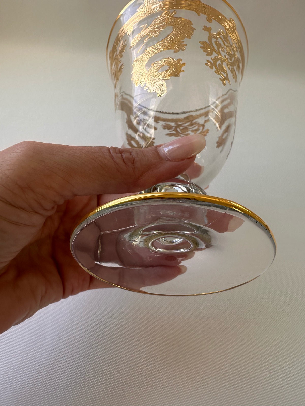 A hand holding a clear glass goblet with ornate gold dragon patterns and a gold rim, photographed against a light neutral background.