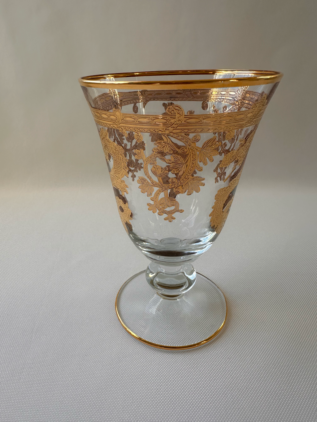 A clear glass goblet with ornate gold scrollwork decoration, featuring a gold rim and a gold-trimmed circular base, shown against a light ivory textured background.