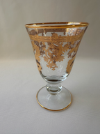 A clear glass goblet with ornate gold scrollwork decoration, featuring a gold rim and a gold-trimmed circular base, shown against a light ivory textured background.