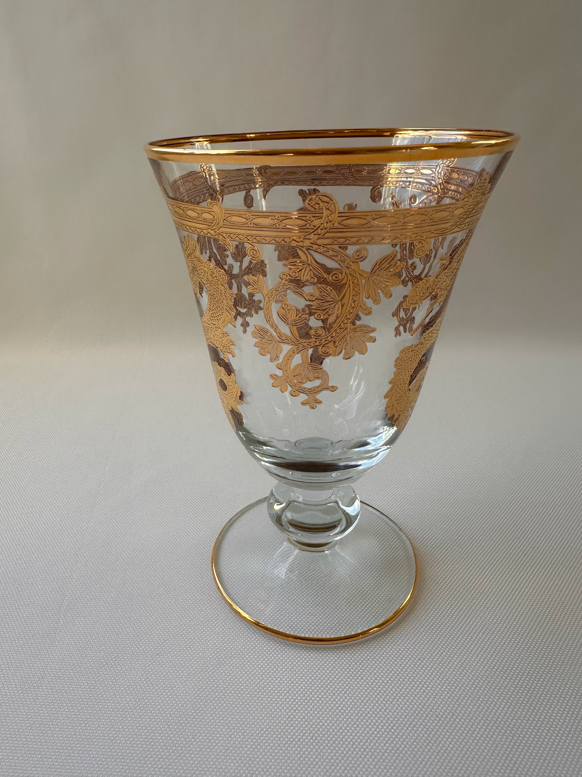 A clear glass goblet with ornate gold scrollwork decoration, featuring a gold rim and a gold-trimmed circular base, shown against a light ivory textured background.