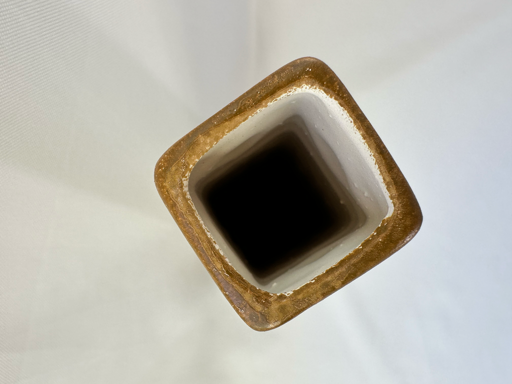 Top-down view of a square ceramic vase with a rustic brown glazed rim and a white interior, shown against a light off-white fabric background.