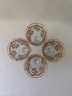 Four clear and gold glass goblets arranged in a flower-like pattern on a plain light background photographed from above, each decorated with a brown dragon motif and gold rim.