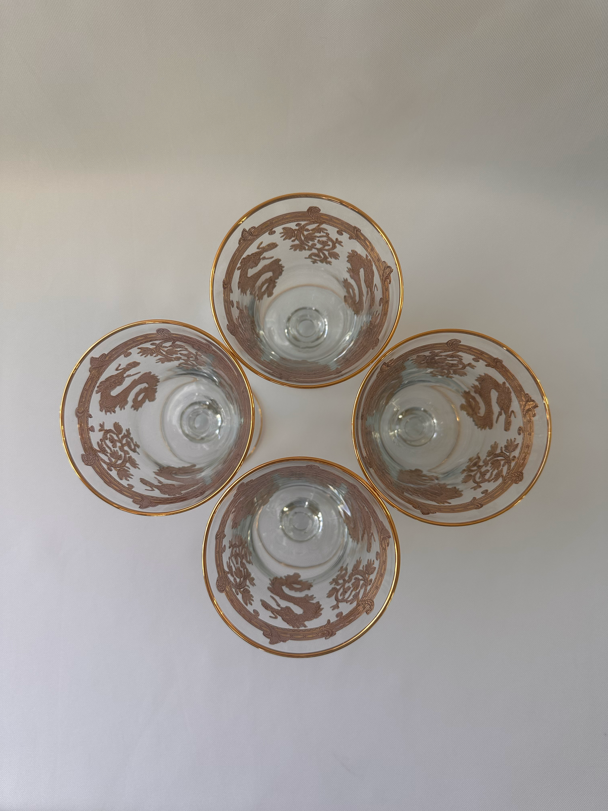 Four clear and gold glass goblets arranged in a flower-like pattern on a plain light background photographed from above, each decorated with a brown dragon motif and gold rim.