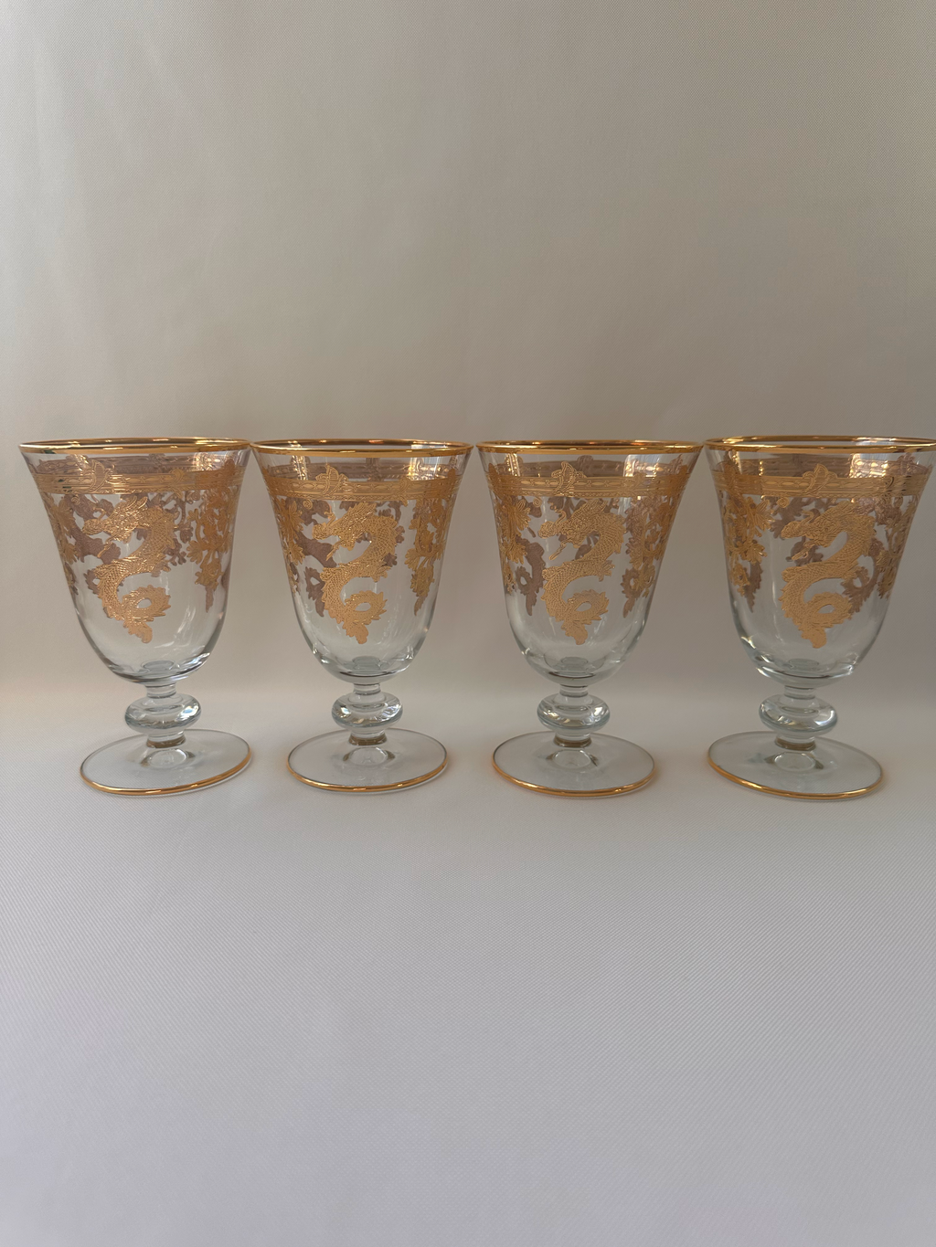 Four clear glass goblets with ornate gold etched patterns and gold rims, arranged vertically on a plain light background.
