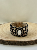 Shell and black beaded statement cuff bracelet displayed on a natural wood slice against a neutral background.

