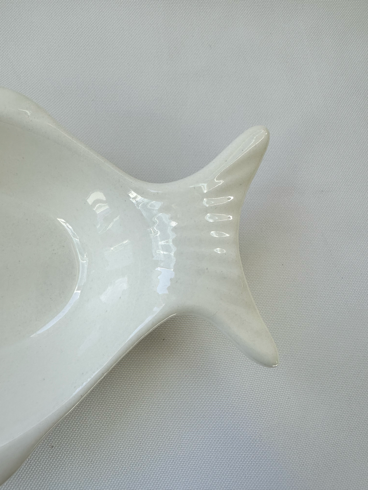 A close-up of a glossy white ceramic dish with a flared, fish-like rim, set against a textured light gray background.