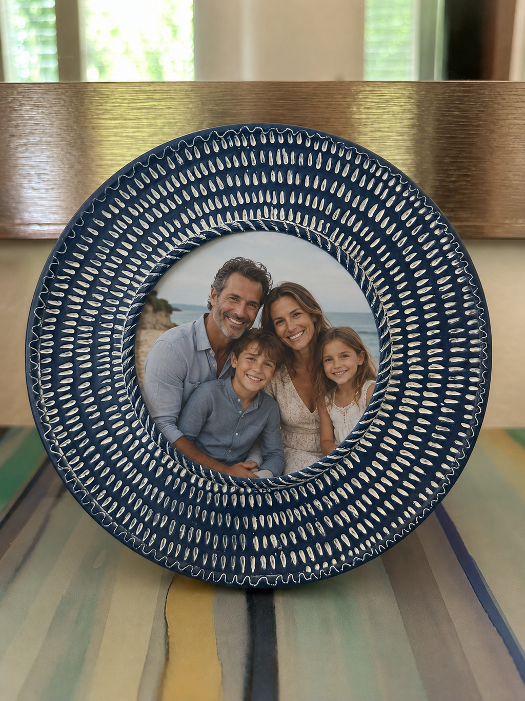 A round Cynthia Rowley decorative picture frame with a textured navy blue and white patterned border, featuring a custom printed family photo in the center, displayed on a decorative box on a console table. 