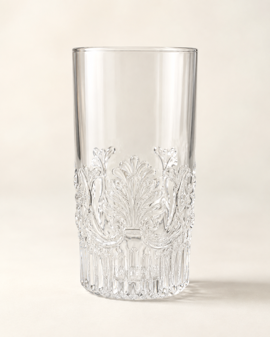 A clear cut-crystal tumbler with ornate floral and scrollwork engravings around the lower half, photographed on a warm off-white background.
