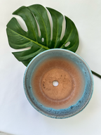 A top-down view of a round ceramic planter with a blue-green glazed rim and a warm terracotta interior, styled with a large green monstera leaf on a white background.